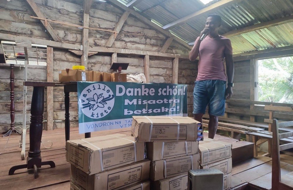 Übergabe im Dorf: Kartons mit Schulheften vor dem Vanillekiste-Banner, ein Sprecher mit Mikrofon